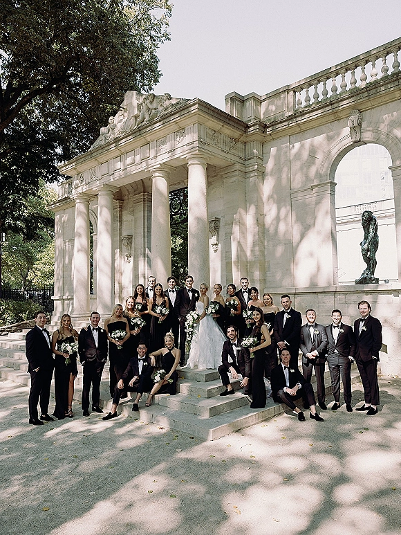 Wedding party portrait with bride centered, bridesmaids and groomsmen in tuxedos and black dresses on stone steps by columned archway, bouquets in hand