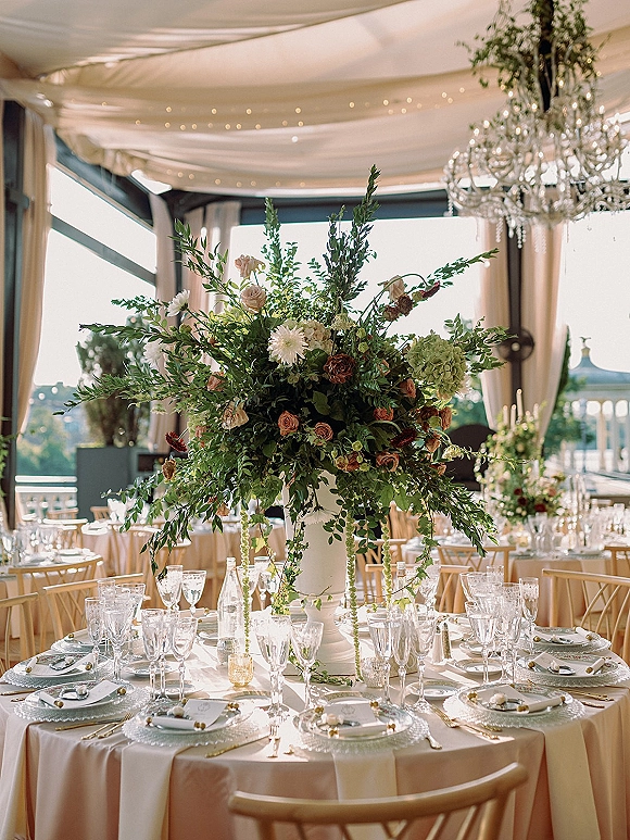 Reception tablescape with wedding table centerpiece in a white urn vase, roses and greenery, under draped tent ceiling and chandelier
