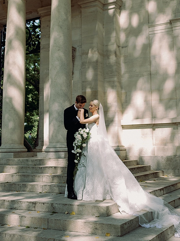 Couple portrait of groom kissing the bride’s hand, her cathedral veil and lace gown flowing over stone steps in dappled light