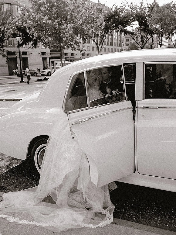Wedding kiss portrait of bride and groom in car, her veil and dress train draped out the door beside a vintage wedding car on a city street