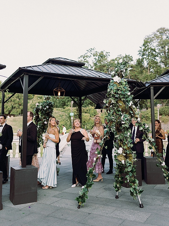 Wedding cocktail hour with wedding guests mingling in cocktail attire, holding drinks under a pergola with hanging lanterns on a garden patio