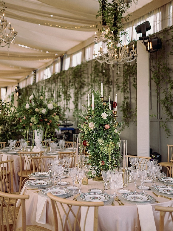 Reception tablescape with a wedding table centerpiece of tall florals, taper candles, and gold flatware under crystal chandeliers in a tent