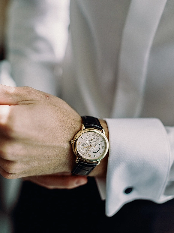 Groom watch detail showing a wedding watch detail with black leather strap, cufflinks on white dress shirt, suit jacket in background