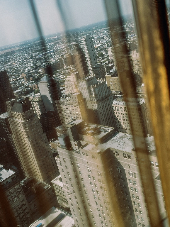 City skyline view framed by window panes and curtains, looking out over downtown skyscrapers and rooftops against a hazy horizon sky
