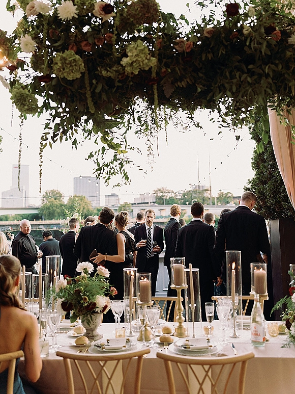 Reception tablescape with outdoor reception tablescape featuring white linens, gold candlesticks, roses, and candles under hanging florals by city skyline