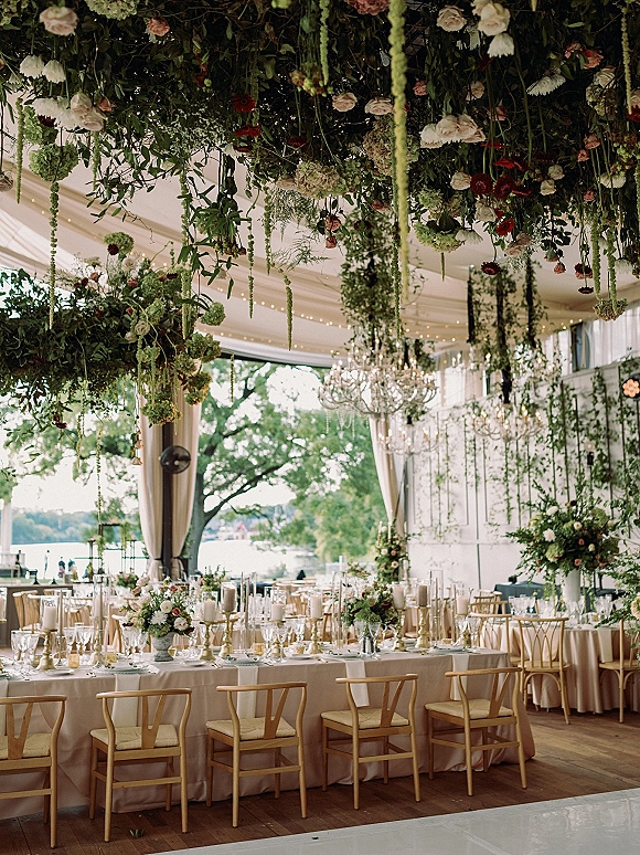 Reception tablescape with head table decor under an overhead floral installation, chandeliers, and candlelit centerpieces in a tented lakefront space