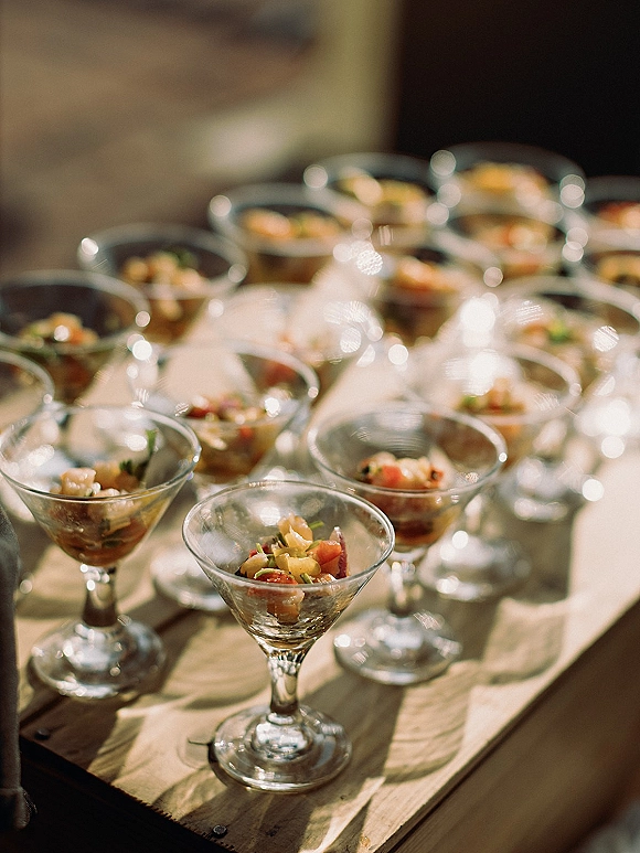 Wedding cocktail appetizers arranged in martini glasses with passed hors d'oeuvres and bite-size hors d'oeuvres on a wooden table, guests blurred behind