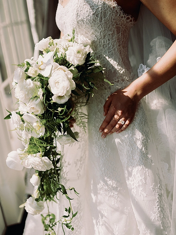 Bridal bouquet with cascading white flowers and greenery held against a lace wedding dress, engagement ring catching soft window light