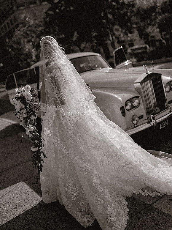 Bridal portrait of a bride with classic car, holding a greenery bouquet in a lace wedding dress and long veil on a tree-lined street