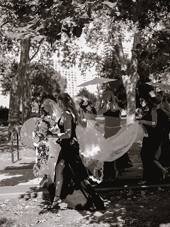 Wedding processional with bride walking down aisle in a long veil, bridesmaids in black dresses holding bouquets on a park walkway under trees