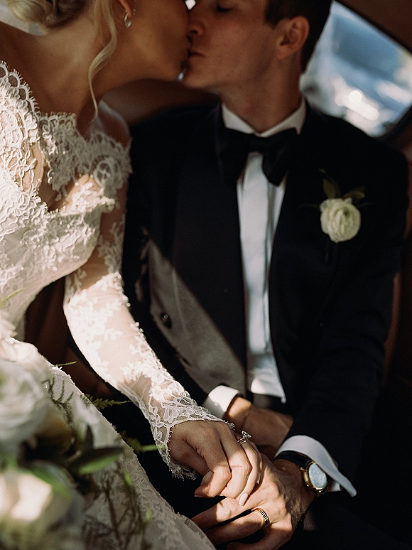Wedding kiss as bride and groom embrace in a car, lace long-sleeve dress and tuxedo with bow tie lit by window light