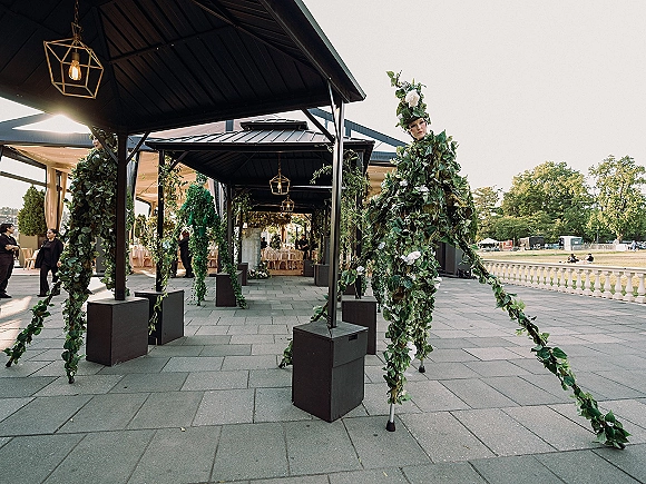 Wedding stilt walkers at cocktail hour in leafy costumes with floral headpieces, towering on a covered patio walkway under hanging lanterns