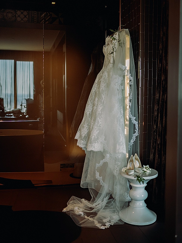 Wedding dress with lace wedding dress bodice hanging by a hotel room window, cathedral veil draped below beside bridal shoes on a side table