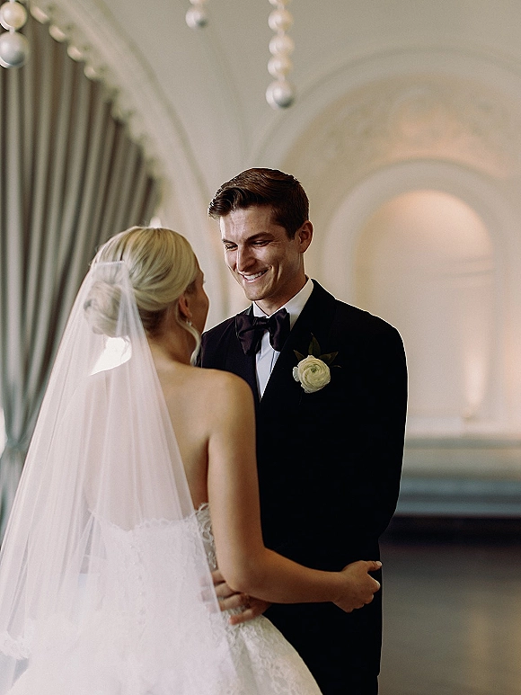 First look moment as bride in veil embraces groom in black tuxedo with rose boutonniere under arched wall and hanging lights indoors