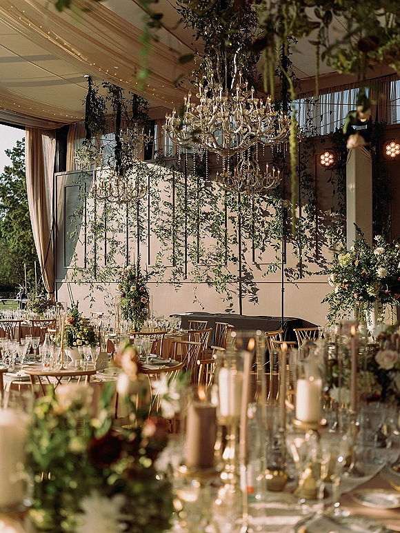 Reception tablescape with crystal chandeliers above round tables, candlelit floral centerpieces, and draped tent ceiling under string lights