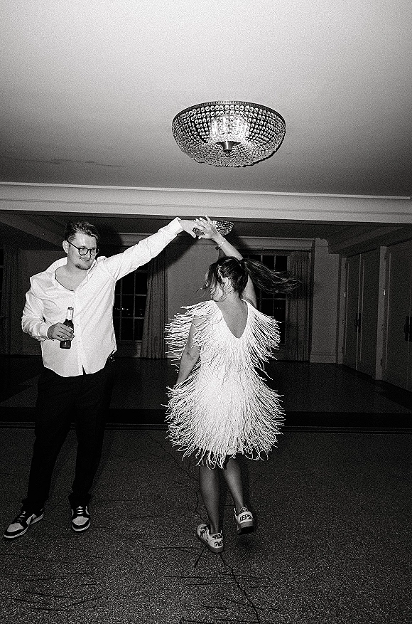 Reception dancing as the couple twirls on the wedding dance floor, bride in fringe dress and sneakers, groom with glasses and beer under a chandelier