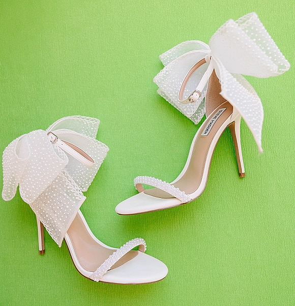 Wedding shoes in white bridal heels with ankle straps, oversized tulle bows, and pearl details arranged on green fabric backdrop