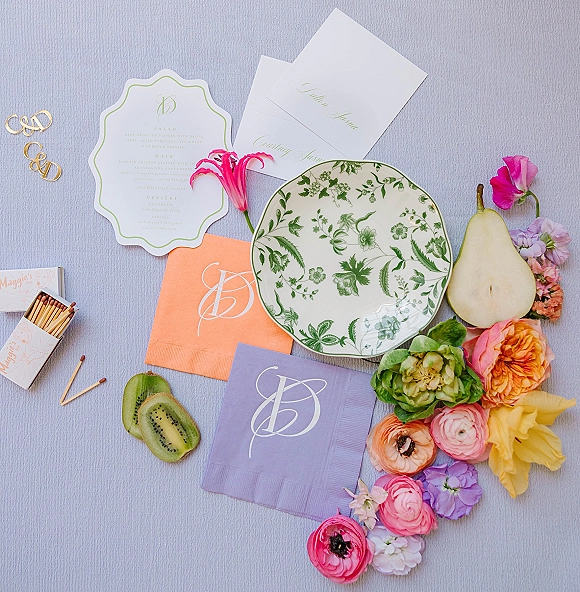 Invitation suite wedding stationery flat lay with menu card, calligraphy envelope, monogram clips, floral plate, fruit, on light blue linen