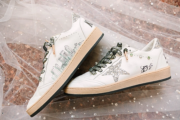 Wedding sneakers with glitter star appliqué and rhinestone details, printed laces and pearl-beaded veil tulle on a pink stone surface