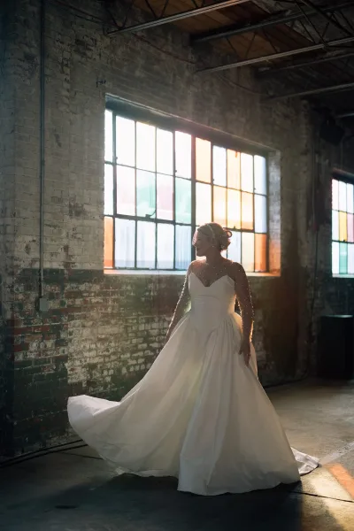 Bridal portrait of a bride in a wedding dress with sleeves, tulle illusion neckline and train, backlit in an industrial loft by stained glass windows