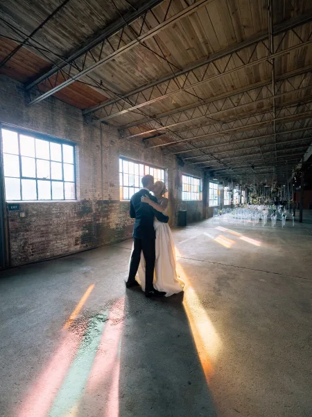 Wedding couple portrait of bride and groom embracing beneath hanging greenery in a brick-walled warehouse with tall factory windows and string lights