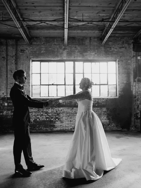 Couple portrait of bride and groom holding hands, facing each other in an industrial loft with brick wall and grid windows, backlit.