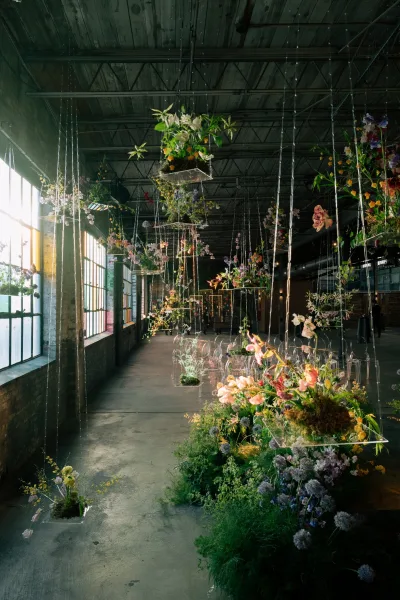 Ceremony aisle decor with hanging floral installation and wildflower arrangements, clear chairs under string lights in an industrial warehouse space