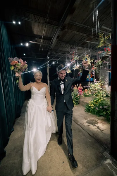 Wedding recessional as bride and groom cheering, holding hands down an industrial warehouse aisle with hanging florals and candles