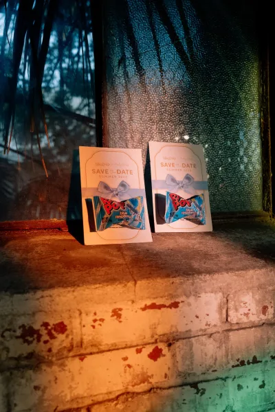 Save the date cards with a ribbon bow and candy favor on a rustic windowsill by a rainy, condensation-streaked window with tree reflections