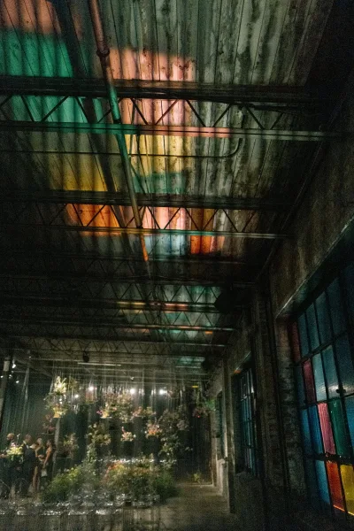 Reception lighting with wedding uplighting casting green and amber glow, string lights and hanging greenery under exposed beams in a warehouse venue
