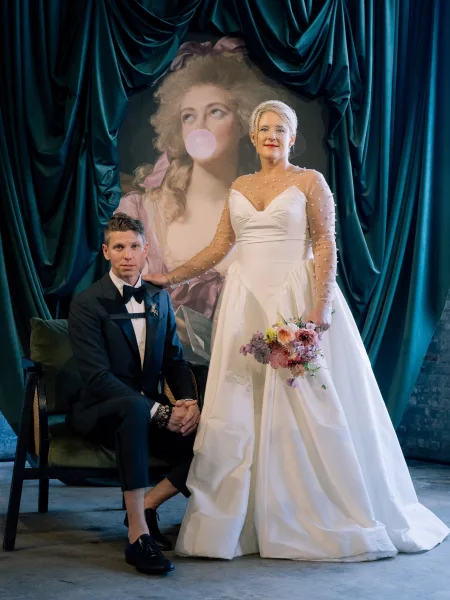 Couple portrait of bride in long sleeve wedding dress and veil beside seated groom in tuxedo, holding bouquet before teal curtain backdrop