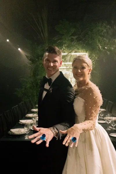 Couple portrait of bride and groom showing rings, bride in pearl sleeve wedding dress beside groom in tux at greenery-lit reception table