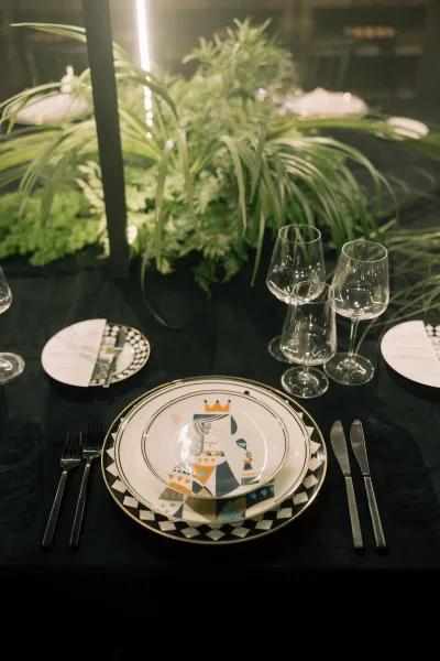 Reception tablescape with wedding place setting on black tablecloth, patterned plates and greenery centerpiece in a dim, moody reception space