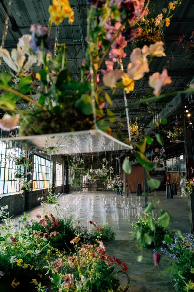 Ceremony setup with hanging florals above clear acrylic chairs in an industrial venue with large factory windows and exposed beams