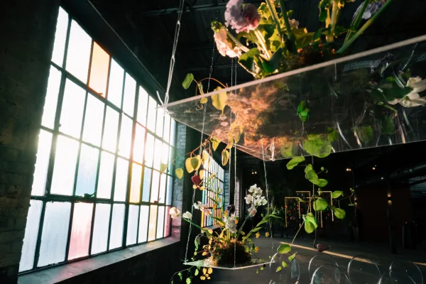Wedding reception centerpiece on an acrylic pedestal centerpiece stand with tall florals, greenery vines, and moss base in an industrial loft by factory windows