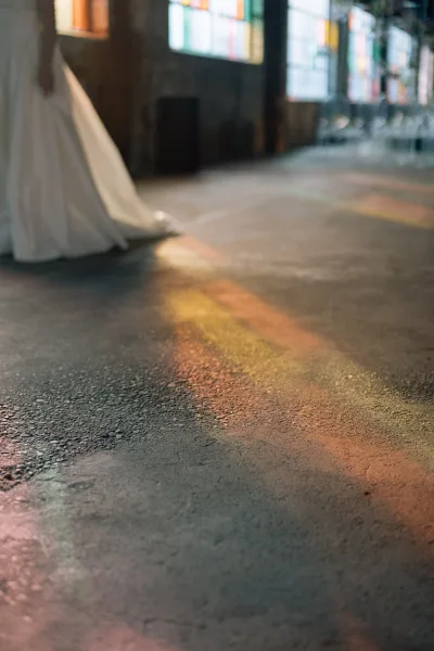 Bridal gown detail with a wedding dress train spread across a concrete floor, lit by stained glass windows in an industrial interior
