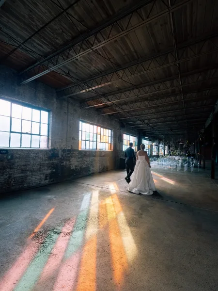 Ceremony moment as bride and groom walk away down the aisle, bridal gown catching rainbow light in an industrial warehouse with brick walls