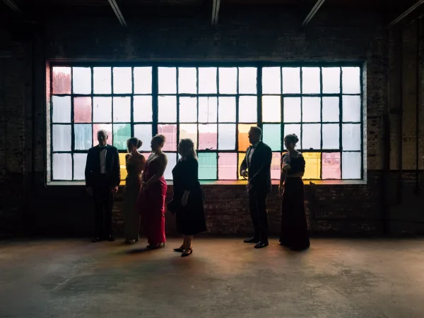 Wedding party portrait of bridesmaids and groomsmen in formal attire, backlit in an industrial loft with brick wall and colorful windows