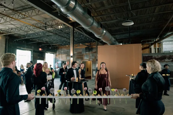 Cocktail hour with wedding cocktail hour guests mingling around high-top tables, passed canapés on trays in an industrial warehouse with string lights