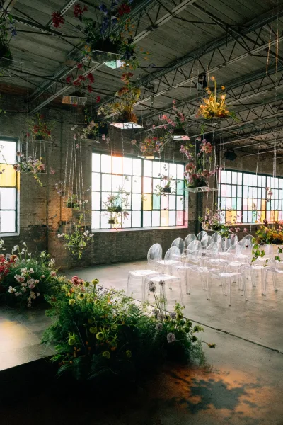 Ceremony setup with hanging florals above clear acrylic chairs, framed by industrial brick walls and large factory windows with stained glass