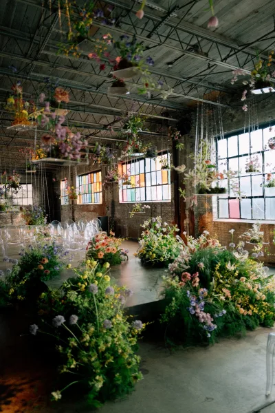 Ceremony setup with indoor ceremony setup featuring meadow-style aisle florals and clear acrylic chairs in an industrial warehouse with grid windows