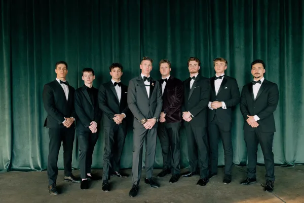 Groomsmen portrait of men in black tuxedos with bow ties and boutonnieres, standing in a lineup before a green velvet curtain indoors