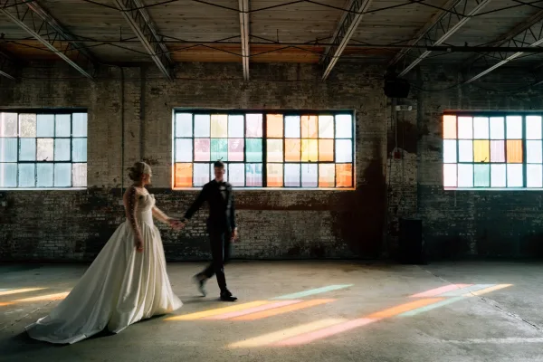 Couple portrait of bride and groom holding hands, walking in an industrial loft with brick wall, colorful window panes, and veil train