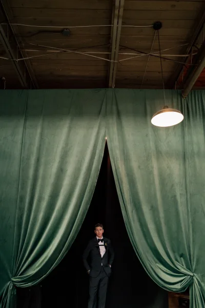 Groom portrait of a black tuxedo groom with boutonniere, hands in pockets, posed by velvet drapes under industrial beams and pendant light
