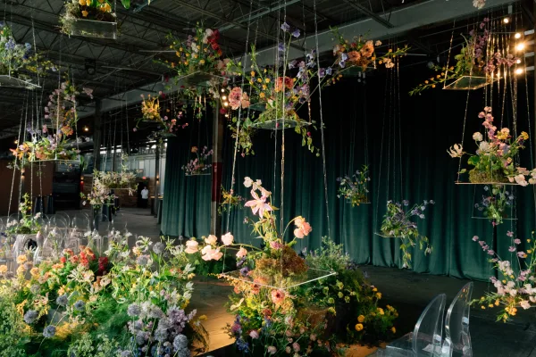 Ceremony backdrop with hanging flower installation, suspended acrylic boxes and greenery, lit by string lights against black draping in an industrial hall