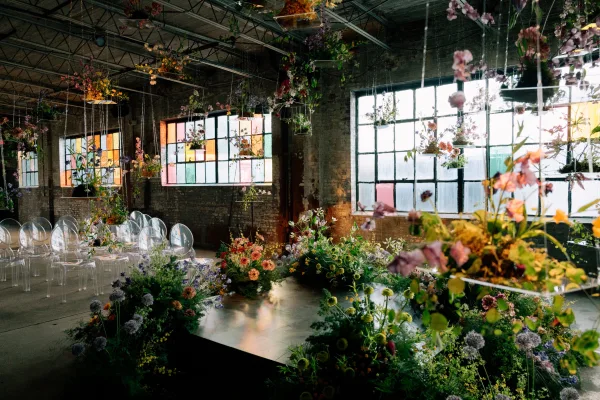Ceremony setup in an industrial wedding ceremony space with clear chairs, hanging floral installation, stained glass panels, and greenery