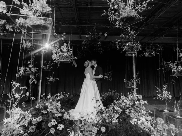 First dance as bride in a gown and groom in tuxedo sway beneath hanging floral installations with stage curtains and overhead lights