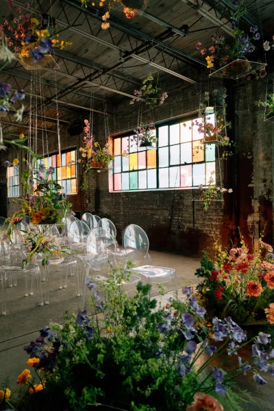 Ceremony setup with indoor ceremony seating on acrylic ghost chairs beneath hanging florals and stained glass windows in an industrial warehouse