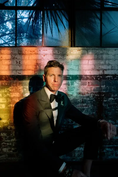 Groom portrait in tuxedo wearing a black bow tie and boutonniere, seated by an industrial brick wall with window light shadows