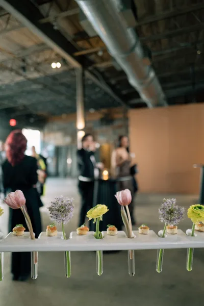 Wedding cocktail hour appetizers on a white display stand with canapes and bud vase stems at an industrial venue with guests behind